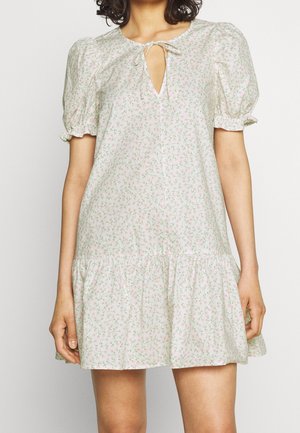 Kvinde iført en kort, lyserød blomsterkjole med pufærmer, flæsekant og rund udskæring bundet med en lille sløjfe.