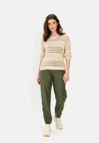 Beżowy sweter robiony na drutach z otwartymi splotami, w połączeniu z oliwkowo-zielonymi spodniami cargo z bocznymi kieszeniami i jasnymi sneakersami. Casualowy, luźny krój.