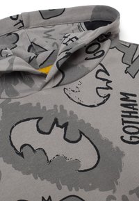 Sudadera gris con un logo negro de Batman y varios patrones gráficos en gris. Presenta una textura suave y un acento amarillo en el interior de la capucha.