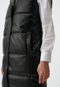 Gilet matelassé en simili cuir noir avec une fermeture zippée, poches latérales et sections rembourrées pour l'isolation. Texture lisse avec une finition brillante.
