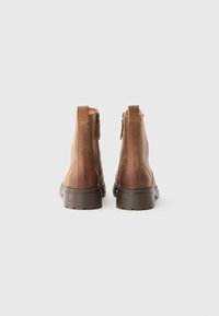 Une paire de bottes brunes en cuir arrivant aux chevilles, vues de l'arrière, avec des semelles épaisses marron foncé et des zips sur les côtés, sur un fond blanc.