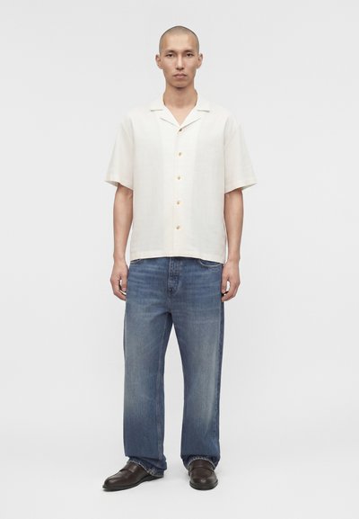 Άνδρας όρθιος, φοράει κρεμ πουκάμισο με κοντό μανίκι και κουμπιά, φαρδιά μπλε τζιν και σκούρα καφέ παπούτσια μπροστά σε απλό γκρι φόντο.