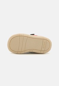 Beige rubber sole with textured pattern and grooves, featuring a black and red upper section with a logo detail at the heel.