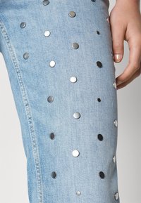 Jean en denim bleu clair avec des détails de clous argentés brillants dispersés et une main reposant doucement sur la cuisse.