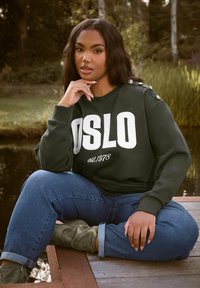 Felpa verde con grafiche "OSLO" e "est. 1978", presenta dettagli a bottone sulle spalle. Abbinata a jeans blu e stivali oliva.