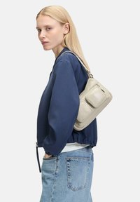 Jasnoszara skórzana torba na ramię z dwiema przednimi kieszeniami, regulowanym paskiem, prostokątnym kształtem i gładką fakturą. Nosi z granatową bluzką i dżinsami.