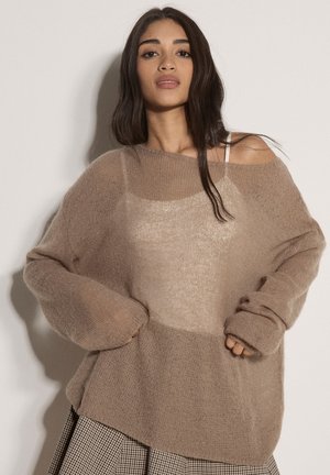 Mujer con suéter suelto y peludo de color marrón claro sobre una blusa beige y falda de cuadros, posando con las manos en los bolsillos del suéter contra un fondo liso.