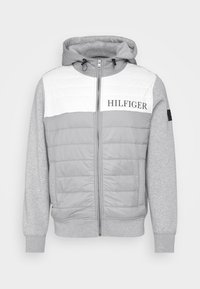 Grijze rits-hoodie gemaakt van een mix van gestructureerde stof en gevoerde delen, met een witte bovenste panel met het "HILFIGER" logo.