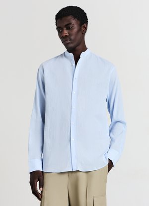 Hellblaues Hemd mit Knöpfen, Mandarin-Kragen, langen Ärmeln und einer strukturierten Stoffqualität. Vorne mit weißen Knöpfen versehen.
