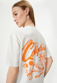 Hvid t-shirt med et orange grafisk design med teksten "Summer Vibe 1998 California" og billeder af palmetræer og bølger på ryggen.