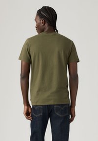 T-shirt verde-oliva de manga curta, feita de algodão, com decote redondo e bainha direita, combinada com calças de ganga azul escuro.