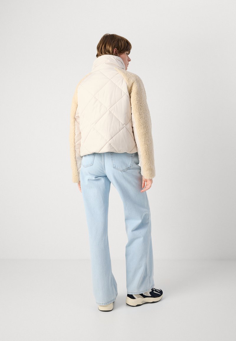 Giacca a sacco con parte anteriore trapuntata beige e maniche in pelliccia color crema, abbinata a jeans a gamba larga blu chiaro e scarpe nere con dettagli bianchi.