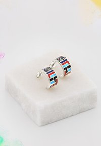 Orecchini a cerchio in argento con motivi geometrici colorati in rosso, blu e nero, esposti su un piedistallo di marmo bianco.