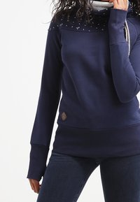 Mörkblå sweatshirt med hög krage, featuring ett mönstrat axelpanel, långa ärmar, och en beige logotyp-patch på nedre sidan.