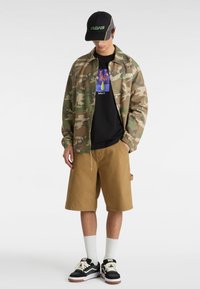 Tarnjacke, schwarzes grafisches T-Shirt, braune Shorts, weiße Socken, schwarz-weiße Sneakers, schwarze Mütze mit grünem Logo. Lässiger Streetwear-Stil.