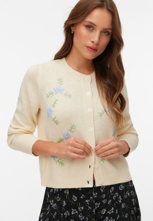 Femme aux longs cheveux bruns portant un cardigan crème avec une broderie florale bleue, boutonnant celui-ci sur une jupe florale noire.
