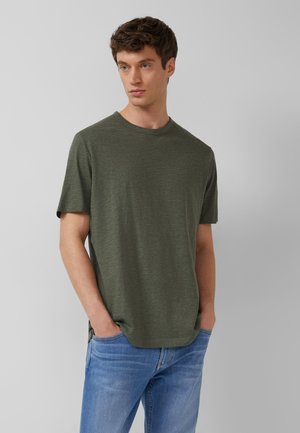 Młody mężczyzna z krótkimi brązowymi włosami, ubrany w oliwkowozielony t-shirt z krótkim rękawem i niebieskie dżinsy, ręce w kieszeniach, patrzy lekko w prawo.