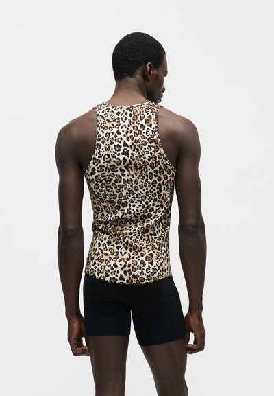 Άνδρας που φοράει αμάνικο τοπ με λεοπάρ σχέδιο και μαύρο σορτς, στέκεται πλάγια με φόντο μονόχρωμο.