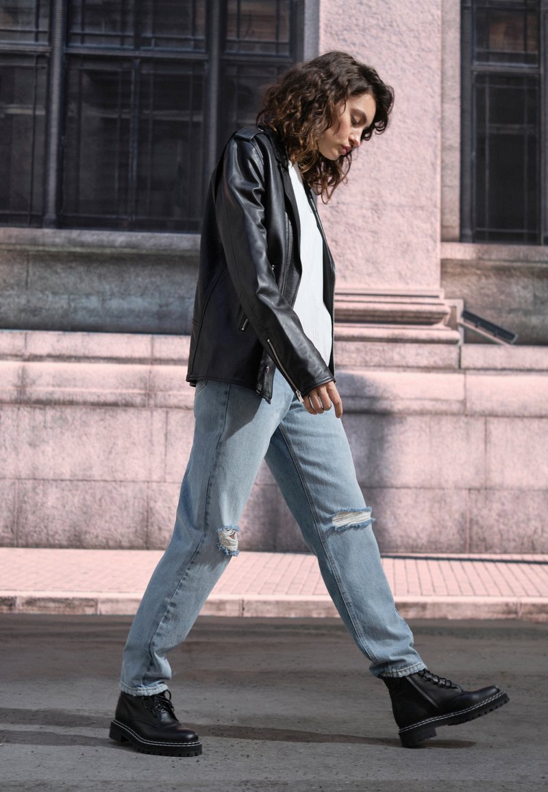 Jaqueta de couro preta, blusa branca, jeans claros desgastados e botas pretas robustas. Ambiente urbano com uma parede de pedra ao fundo.