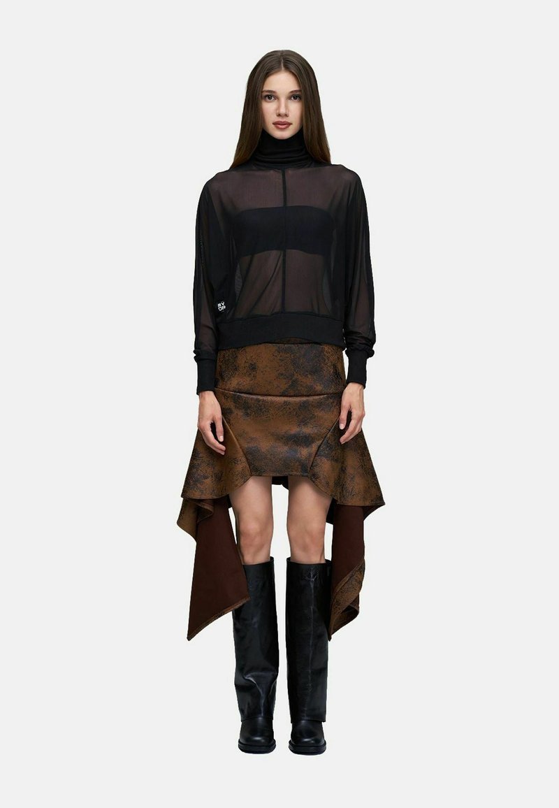 Femme aux longs cheveux bruns portant un haut noir transparent à col roulé, une jupe asymétrique marron et des bottes noires montant jusqu'aux genoux, debout sur un fond blanc.