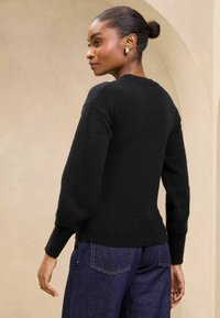 Maglione nero lavorato a maglia con scollo tondo, orlo e polsini a costine, abbinato a jeans in denim scuro, visto da dietro. Texture liscia.