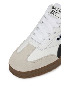 Białe skórzane sneakersy z beżowymi zamszowymi akcentami, czarnym brandingiem i brązową gumową podeszwą. Posiadają białe sznurowadła i wyściełany język.