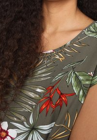 Hombros y parte superior del pecho de una persona que lleva una camiseta verde con estampado de hojas tropicales y flores rojas, y cabello rizado de color marrón oscuro.
