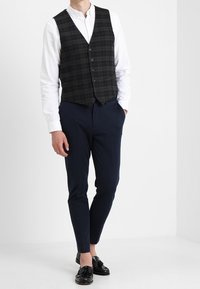 Ciemnozielona kamizelka w kratę z pięcioma guzikami, noszona na białej koszuli, w połączeniu z granatowymi spodniami i czarnymi butami. Prosty design z dopasowanym krojem.