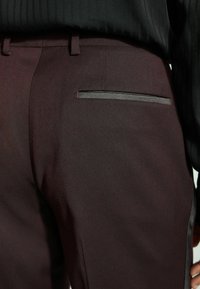 Burgundinės kelnės su lygiu paviršiumi, su stilingu juodu akcentu ant užpakalinės kišenės ir pritaikytu siluetu.