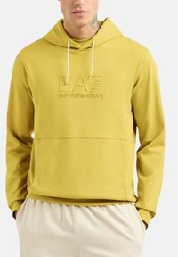 Uomo che indossa un hoodie color senape di Emporio Armani con logo in rilievo e pantaloni avorio, mani in tasca, tatuaggio visibile sul collo.