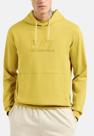 Homme portant un hoodie couleur moutarde de Emporio Armani avec un logo en relief et un pantalon crème, les mains dans les poches, un tatouage visible sur le cou.