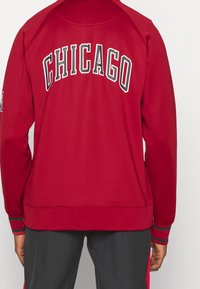 Rød sweatshirt med "CHICAGO" i fed sort og hvid skrift på ryggen. Ribbet manchetter og krave med en struktureret stofoverflade.