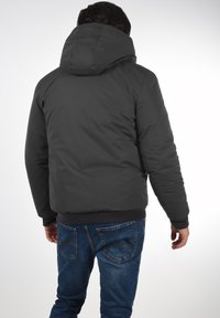 Mørk grå dunjakke med hætte, der har elastiske manchetter og ribbet talje. Båret over blå jeans med synlige baglommer.