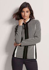Schwarze und weiße gemusterte Jacke aus strukturiertem Stoff, hochgeschlossen, mit Frontreißverschluss und kontrastierenden vertikalen schwarzen und weißen Streifen an den Seiten.