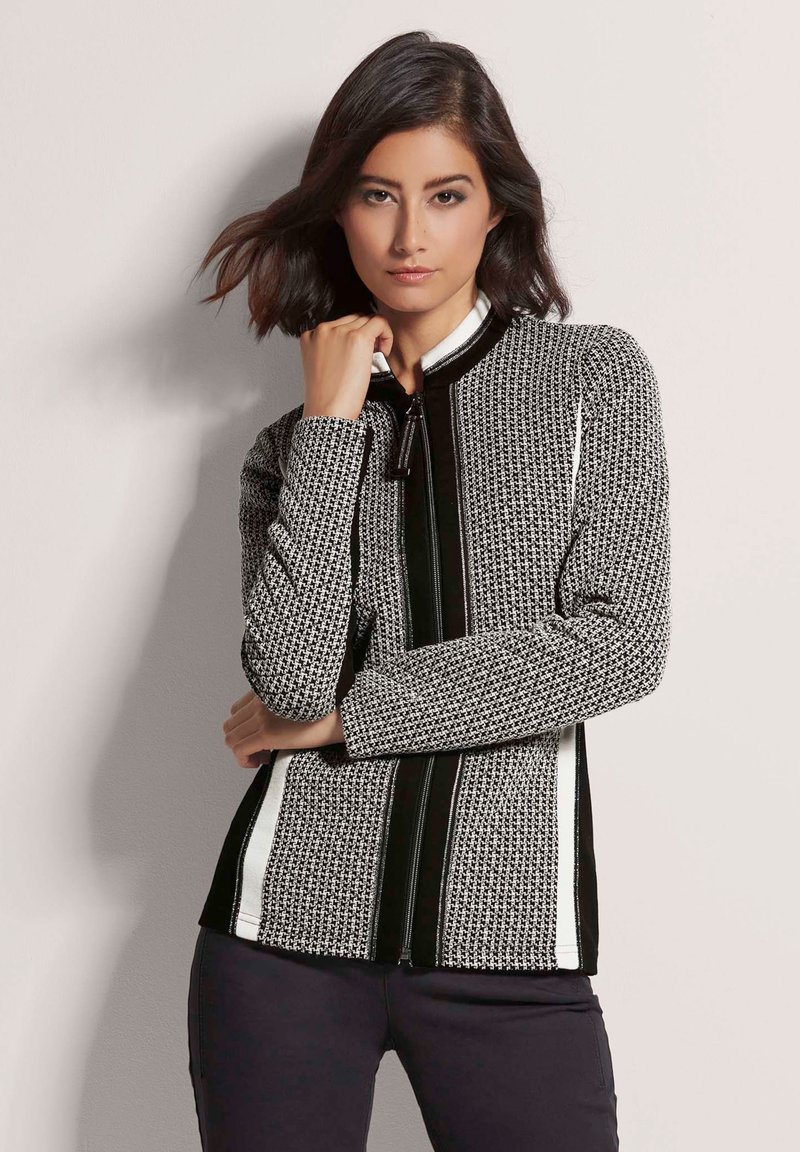 Schwarze und weiße gemusterte Jacke aus strukturiertem Stoff, hochgeschlossen, mit Frontreißverschluss und kontrastierenden vertikalen schwarzen und weißen Streifen an den Seiten.