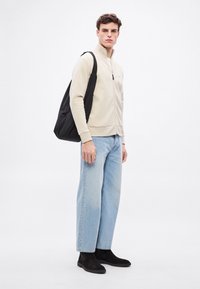 Bézs cipzáras pulóver, világoskék bő nadrág, fekete szarvasbőr bokacsizma és fekete válltáska. Simább anyag és laza fazon.