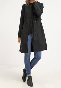 Schwarzer langer Mantel aus weichem Stoff, mit geradem Schnitt, fünf Knöpfen und kragenlosem Design, kombiniert mit blauen Jeans und schwarzen Stiefeln.