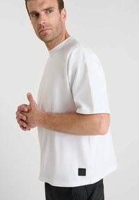 T-shirt bianco a maniche corte con scollatura a girocollo, vestibilità rilassata e un piccolo etichetta nera nella parte bassa della schiena, realizzato in un tessuto liscio e leggero.
