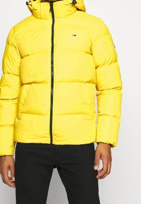 Žlutá péřová bunda se zipem vpředu, vysokým límcem a kapucí. Má prošívanou strukturu a boční kapsy. Logo na hrudi. Černé kalhoty.