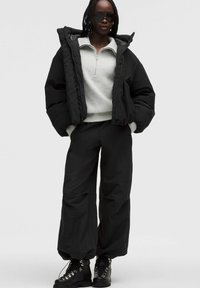 Sort puffetjakke med høj krave, lysegrå sweater og løse sorte bukser. Tunge sorte snørebodder fuldender outfittet.