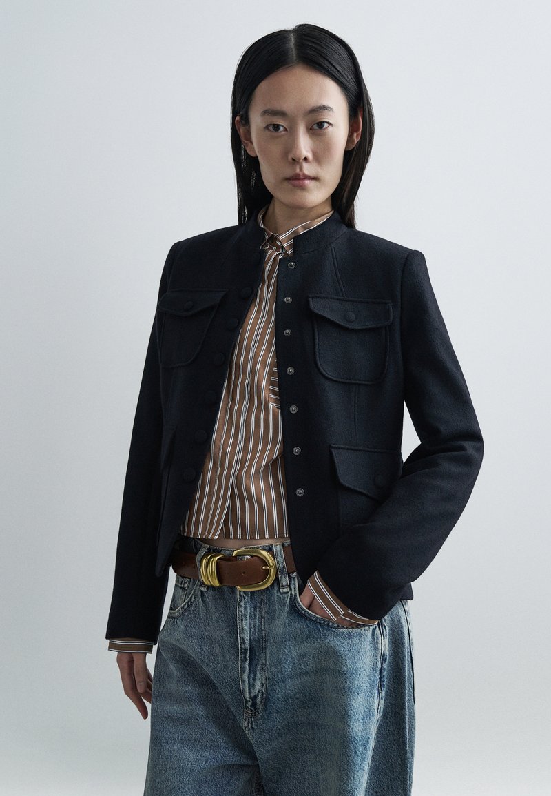 Sort tætsiddende jakke med to brystlommer, kombineret med en stribet brun skjorte og højtailede blå jeans, med et brunt bælte med guldlås.