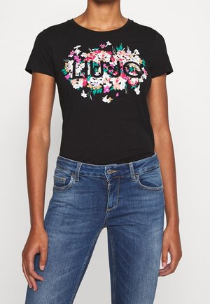 T-shirt noir à manches courtes avec un motif floral coloré et le texte "LIUJO", porté avec un jean bleu sur une personne debout.