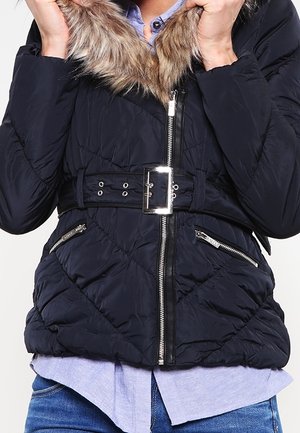 Person iført en navyquiltet jakke med krave foret med pels, frontlynlås, bælte med sølvspænde og lynlåslommer i siden, kombineret med blå jeans.
