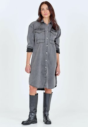 Vrouw met lang bruin haar, gekleed in een grijze denimjurk met knopen en opgerolde mouwen en zwarte kniehoge laarzen, staand tegen een witte achtergrond.