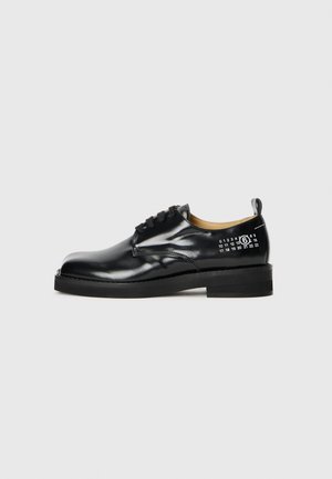 Zapatos de cuero negro con cordones, acabado liso, punta redondeada y una silueta estructurada. Presentan detalles numéricos en blanco en el lateral.