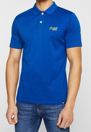 Mand iført en kongeblå kortærmet poloshirt med to knapper og lille mærkelogo på brystet, kombineret med blå jeans.