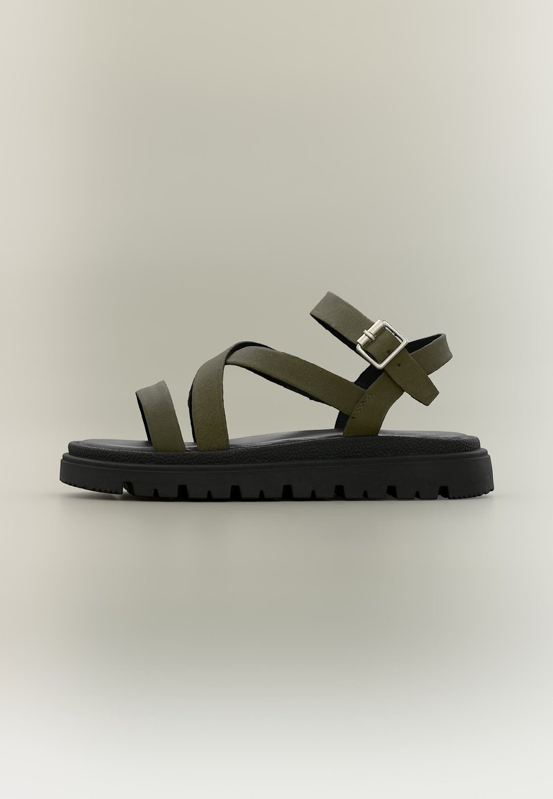 Πράσινο σανδάλι με λουράκια και μαύρη ανθεκτική σόλα, με ασημένιο κούμπωμα, σε ουδέτερο φόντο.
