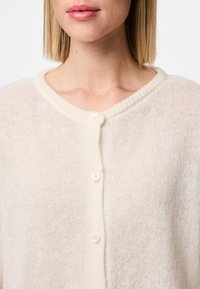 Cream-colored knit cardigan with a ribbed neckline, featuring five buttons and a lightweight, textured fabric.