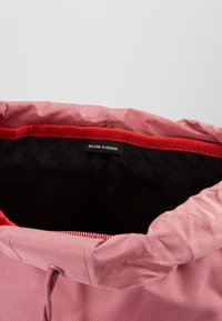 Sac en tissu rose avec une doublure intérieure rouge et un intérieur en polaire noire. Dispose d'une fermeture à cordon et d'une étiquette indiquant "BIENVENUE CHEZ HERSCHEL."