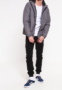 Man som bär grå jacka, vit t-shirt, svarta cargo-byxor, vita strumpor och blå Adidas-sneakers, står mot vit bakgrund.